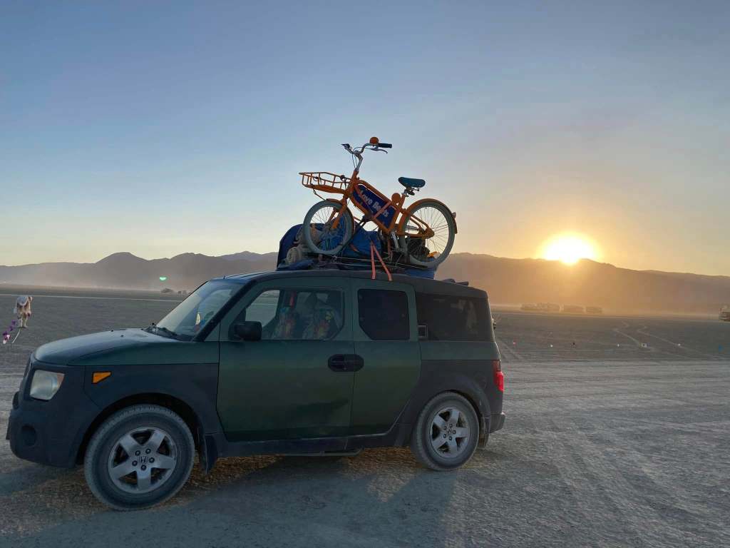 A green SUV parked on a dusty landscape with a bicycle strapped on the roof, set against a sunset backdrop.