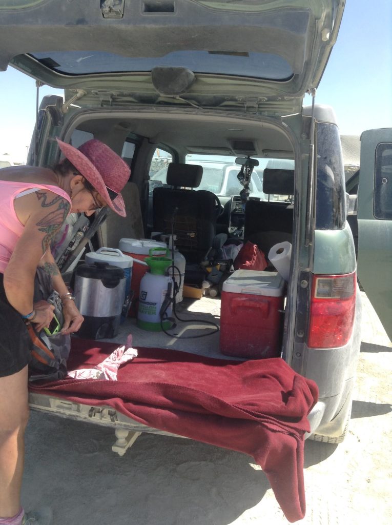 A woman wearing a pink hat and tank top is organizing items in the back of a parked vehicle. The vehicle contains various supplies, including a cooler, a blanket, and cleaning tools, in a dusty outdoor setting.
