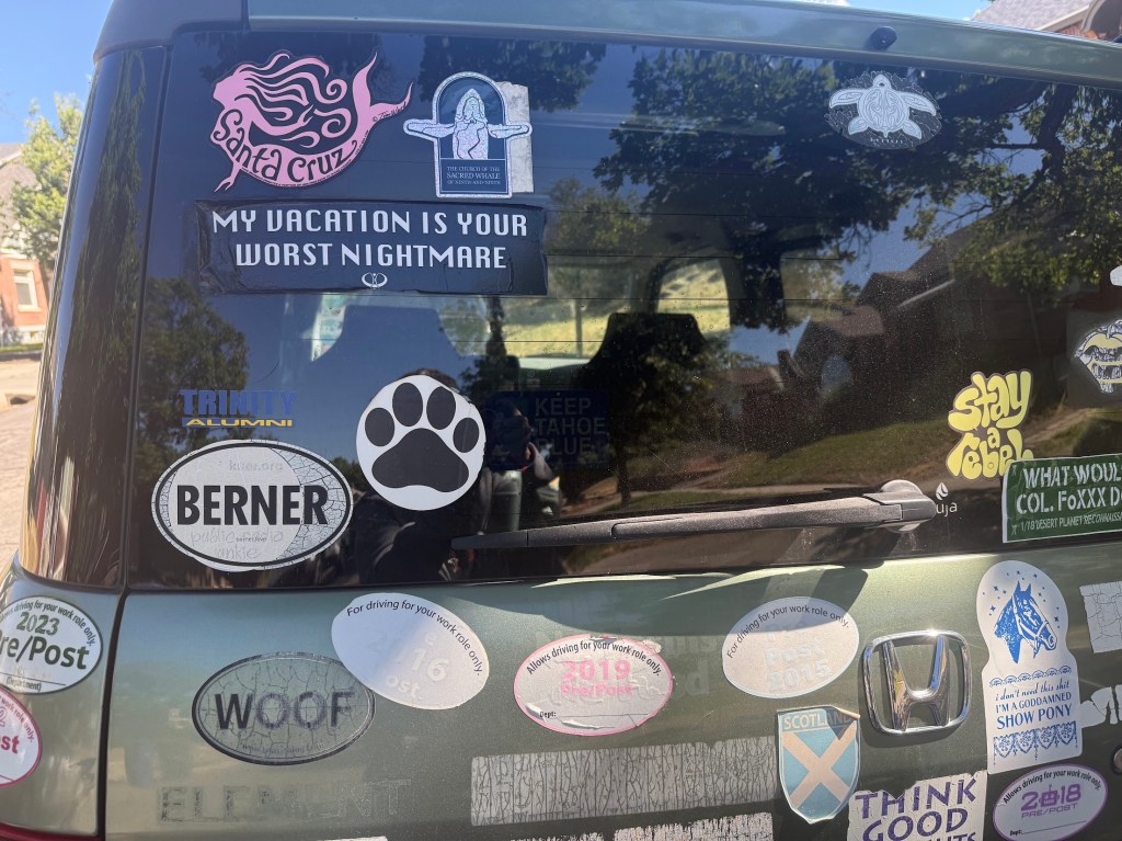 The back of a vehicle covered in various bumper stickers, including one that reads 'My vacation is your worst nightmare' and several others promoting places and organizations like 'Santa Cruz', 'Trinity Alumni', and dog-related stickers.