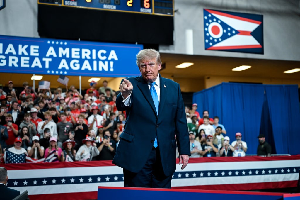 Donald Trump gestures towards the camera during a rally, with a crowd of supporters in the background and a banner reading 'MAKE AMERICA GREAT AGAIN!'